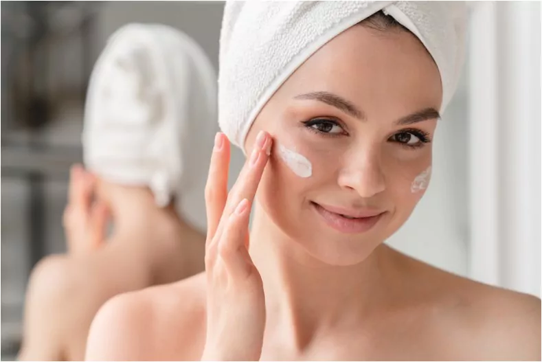 young beautiful woman in bath towel smiling applying moisturiser cream on her face