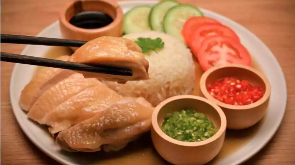 hainanese chicken rice served with fresh vegetables and special sauces on wooden table