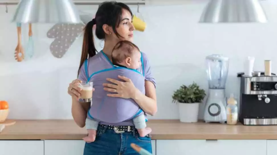 shot of pretty young mother with little baby in sling drinking coffee in the kitchen at home