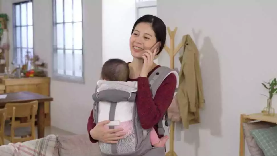 waist up asian woman wearing a baby carrier is laughing during a phone talk while her newborn girl is cradled in her arms at home