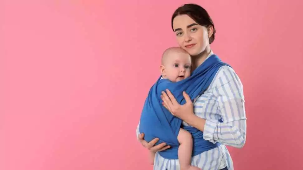 mother holding her child in sling (baby carrier) on pink background