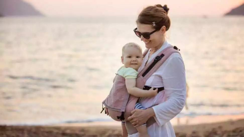 cheerful caucasian woman with baby daughter in buckle carrier on beach at sunset