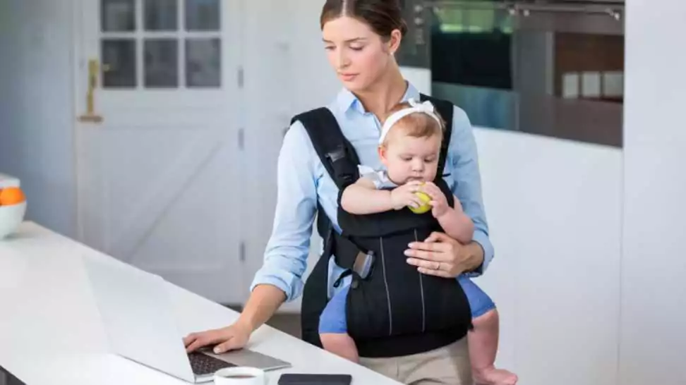 woman carrying baby girl while using laptop at table in house