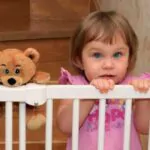 little blonde girl on the stairs with a gate with a teddy bear friend