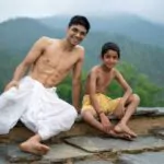 a young man sitting on the roof with his younger brother both wearing dhoti smiling into the camera