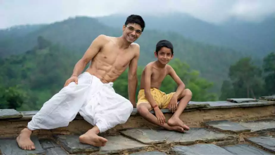 a young man sitting on the roof with his younger brother both wearing dhoti smiling into the camera
