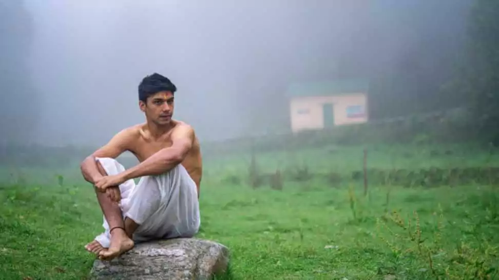 young man wearing a dhoti sitting on a rock open in the green field in a foggy morning