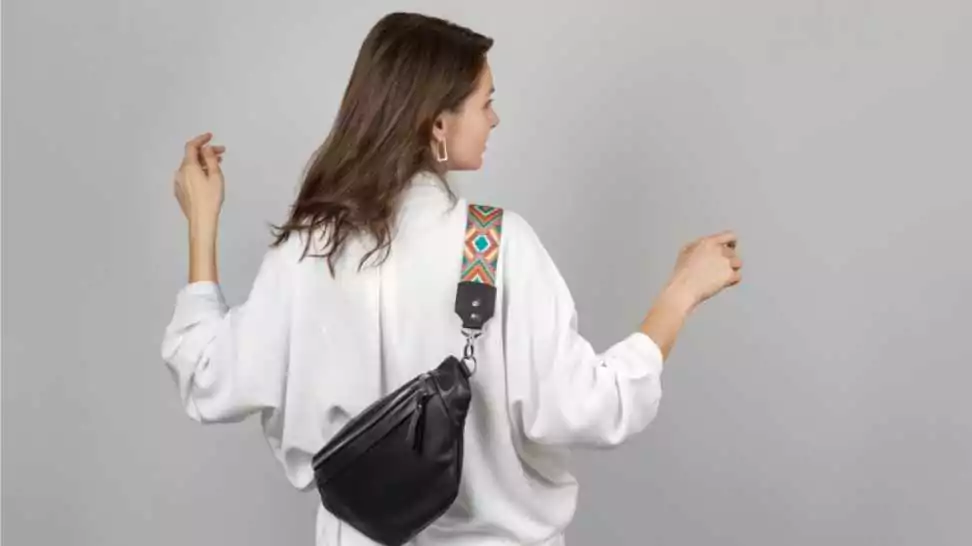 white woman wearing white jeans shirt and crossbody black leather belt bag over grey background
