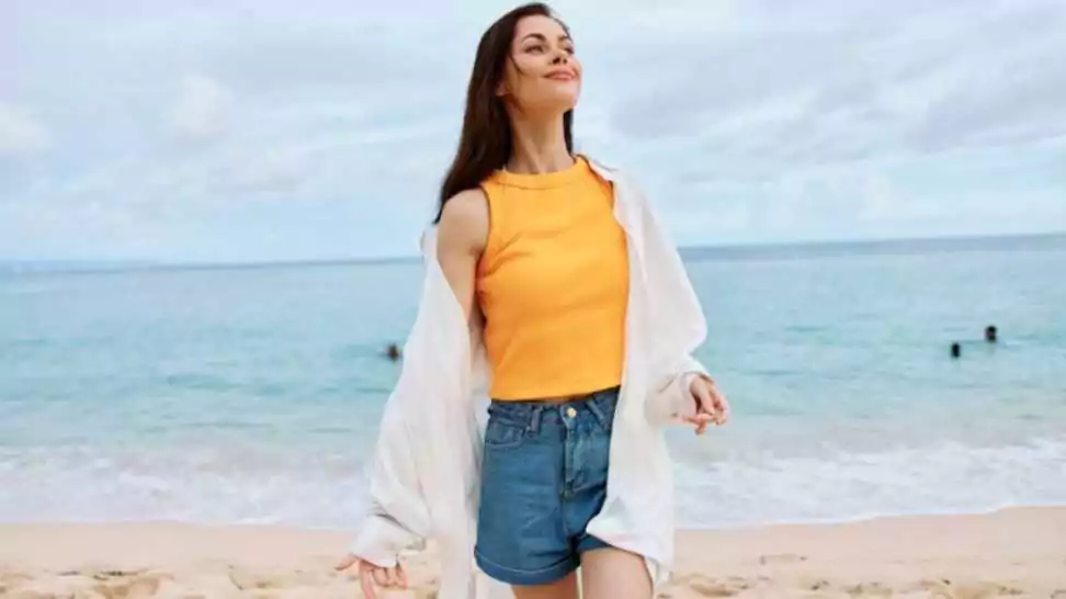 woman with long hair brunette walks along the beach in a yellow tank top denim shorts and a white shirt by the sea summer