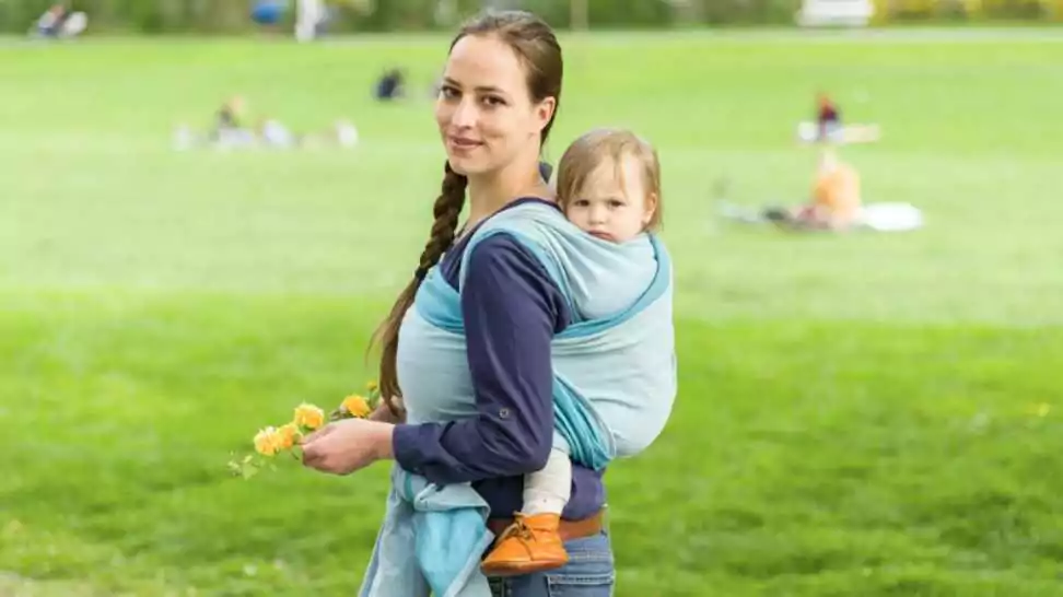 a young mother carrying her little baby in a sling on her back and walked in beautiful sunny weather by a park