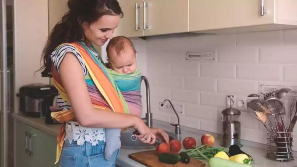 young mom cooking in the kitchen with the little baby in a sling