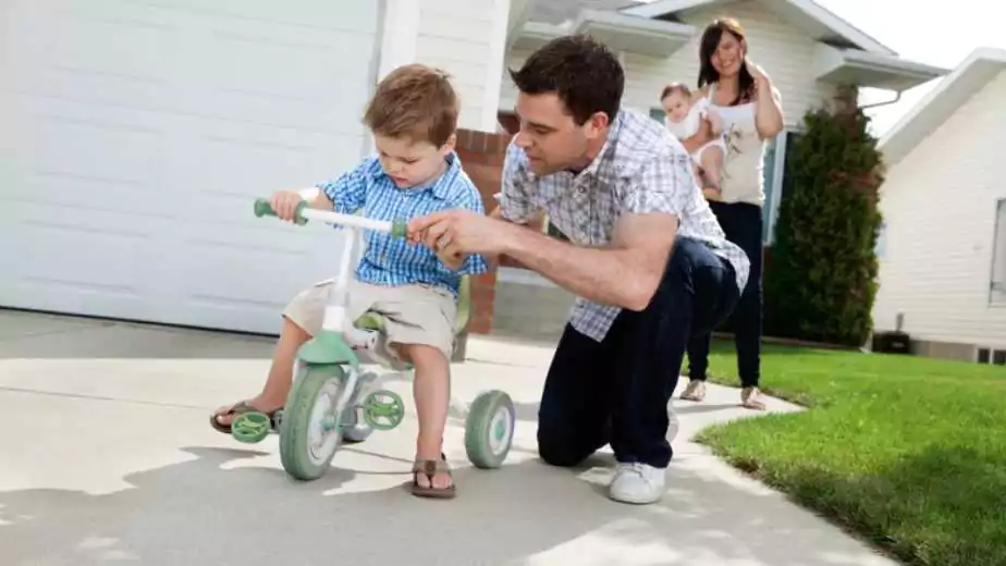 father teaching his son to ride tricycle while wife standing in background