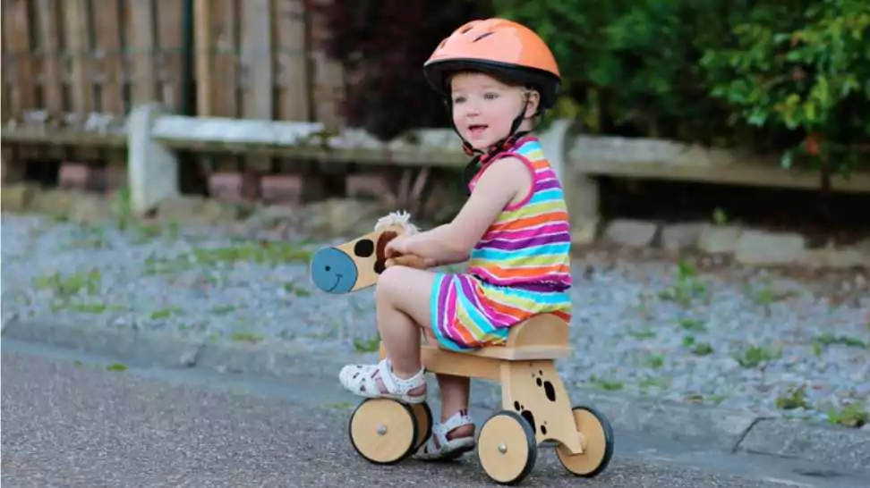 toddler girl in colorful dress and orange safety helmet playing outdoors on the street learning to balance on her three wheel bike