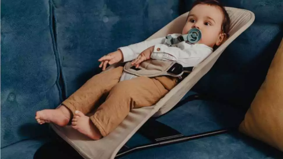cute and joyful baby boy with pacifier and bow tie sitting in a bouncer next to a window and watching cartoons