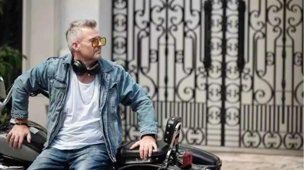 cool stylish mature man in jeans and denim jacket leaning on motorcycle and looking away