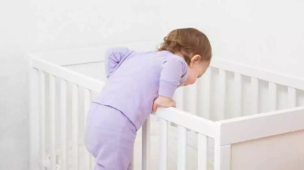 baby girl in purple pyjama trying to get in white crib and going to sleep