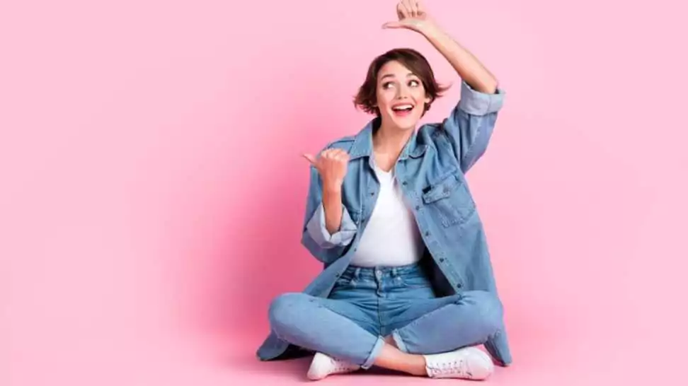 full length photo of charming dreamy lady dressed denim jacket legs crossed thumbs empty space isolated pink color background