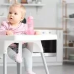 cute baby girl with bottle of water sitting on high chair in kitchen