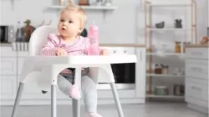 cute baby girl with bottle of water sitting on high chair in kitchen