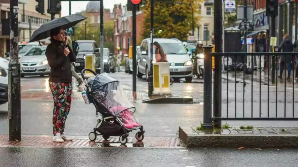 mother with her baby in an umbrella stroller sheltered under umbrella during the rain is crossing the street