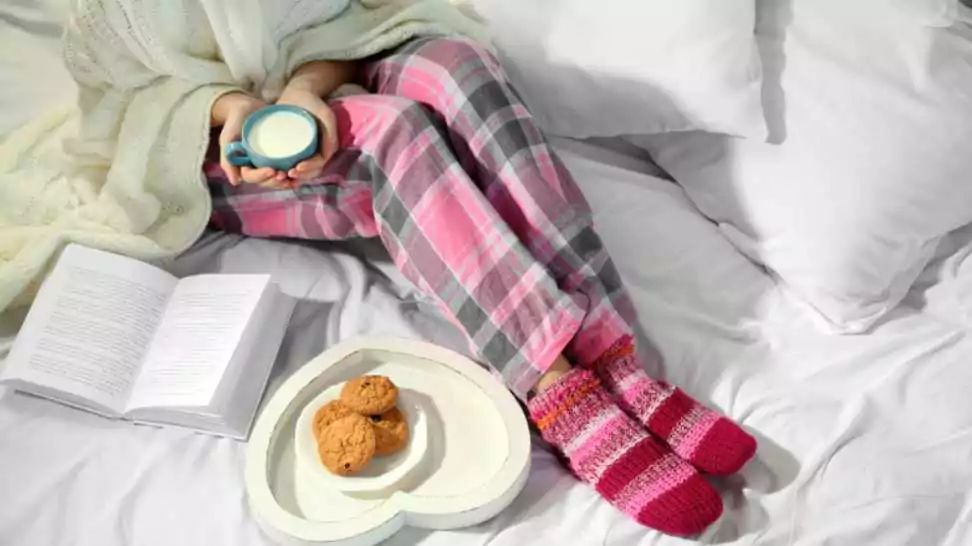 woman in pajamas reading a book and drinking milk on her bed
