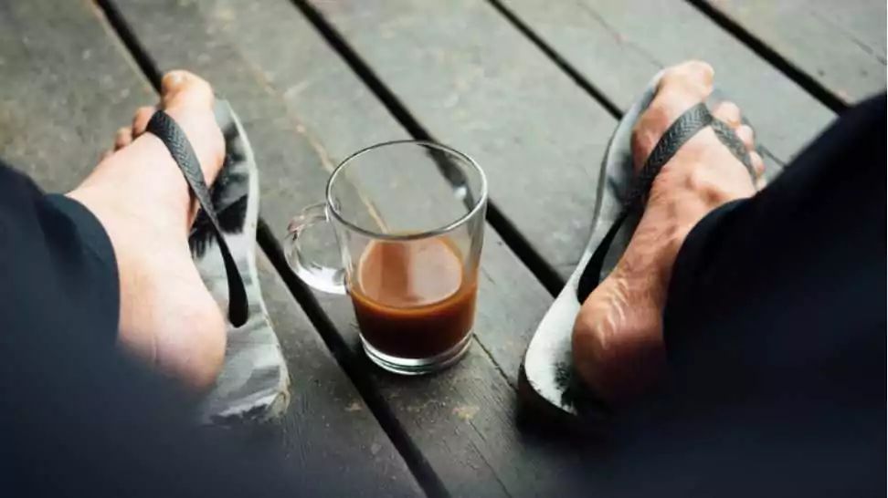 the feets of a man wearing black and white flip flops or thongs on a outdoor of wood plank flooring and with a cup of coffee on the floor