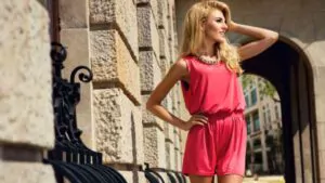 beautiful blonde woman wearing pink jumpsuit and black sunglasses posing on the wall