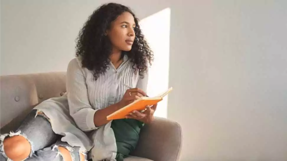 attractive young dark skinned female with afro hairstyle relaxing on couch at home having pensive thoughtful look writing down ideas for her own startup project