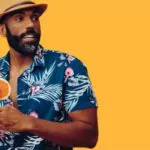 handsome bearded mid adult african american man wearing hawaiian floral shirt and hat smiling with orange juice cocktail