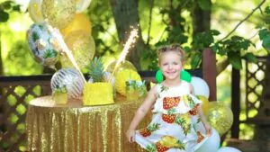 little beautiful girl with pigtails and in a dress with pineapples for a birthday next to it stands a festively decorated table with a delicious pineapple cake